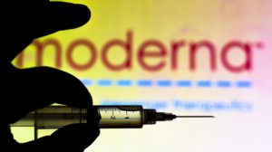 the medical syringe (coronavirus vaccine) is seen with Moderna Therapeutics company logo displayed on a screen in the background-Coronavirus Vaccine News-ss-featured