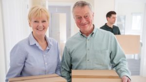 Portrait Of Senior Couple Downsizing In Retirement Carrying Boxes Into New Home On Moving Day With Removal Man Helping | home downsizing tips