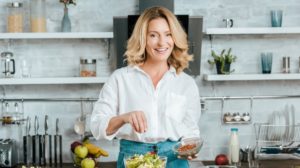 beautiful adult woman spilling spice onto lettuce in bowl at kitchen and looking at camera | Diet For Women Over 60 | Elements Of Healthy Eating | Featured