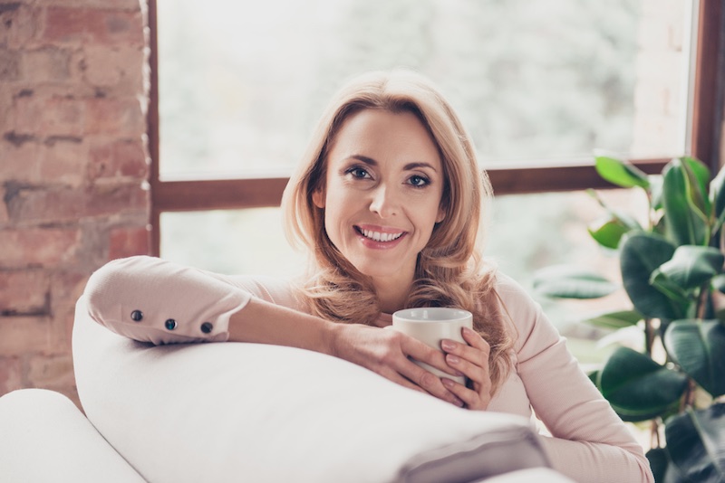Close up portrait of smiling happy charming beautiful dreamy with blonde curly hair mature woman, she is drinking fresh morning coffee and sitting on a sofa at home