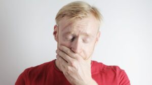 stressed male worker covering mouth with hand against gray background | How To Get Rid Of Bad Breath & How to Prevent It | Featured