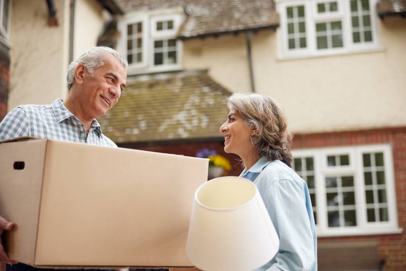 Mature Couple Carrying Boxes On Moving Day In Front Of Dream Home | Downsizing for Retirement