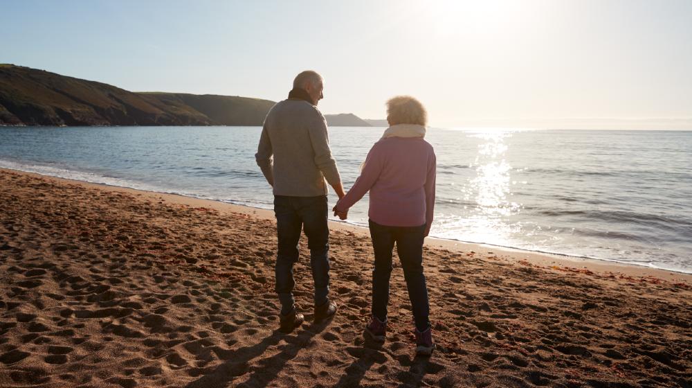 Rear View Of Loving Retired Couple Holding Hands Looking Out To Sea On Winter Beach Vacation | Downsizing for Retirement | featured
