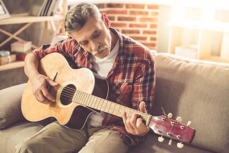 Handsome mature man in casual clothes is smiling while playing guitar at home | growing old