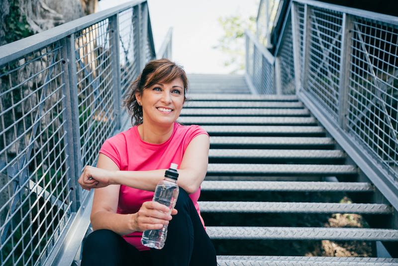 Happy and beautiful middle aged woman sitting on metallic stairs relaxing before running outdoors holding a water bottle | Amazing Benefits Of Turning 50
