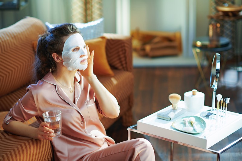middle age woman in pajamas with white sheet facial mask on face and glass of water in the modern living room in sunny winter day. | making lifestyle changes