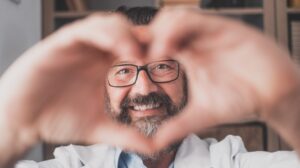 Close up portrait of smiling young caucasian male nurse or GP in white medical uniform show heart love hand gesture | Why Good Nutrition and Physical Fitness Is Vital to Good Health | featured