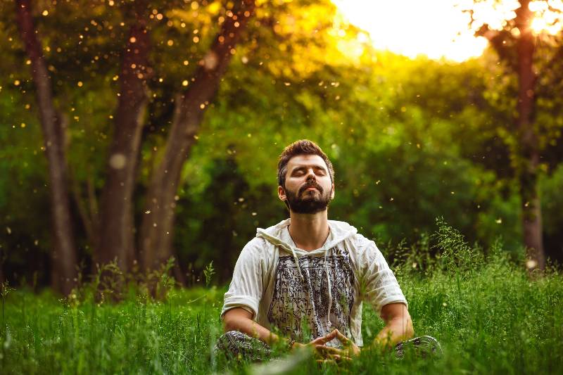 A bearded man is meditating on green grass in the park with face raised up to sky and eyes closed-COVID Immunity