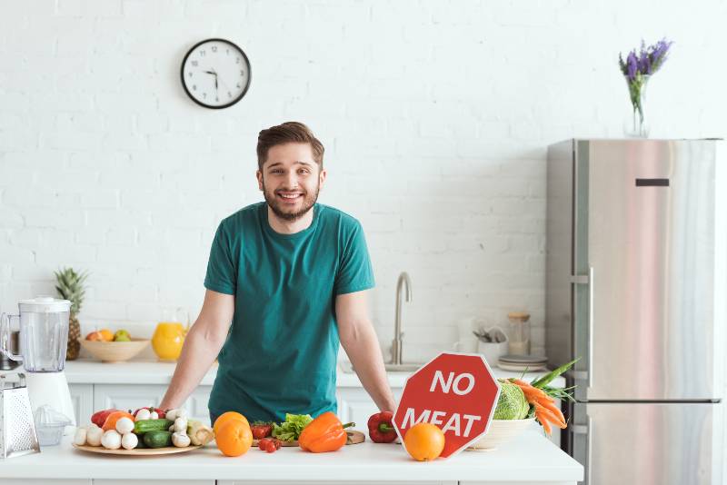 smiling handsome vegan man standing near kitchen counter with vegetables and no meat sign-what can you eat on the daniel fast