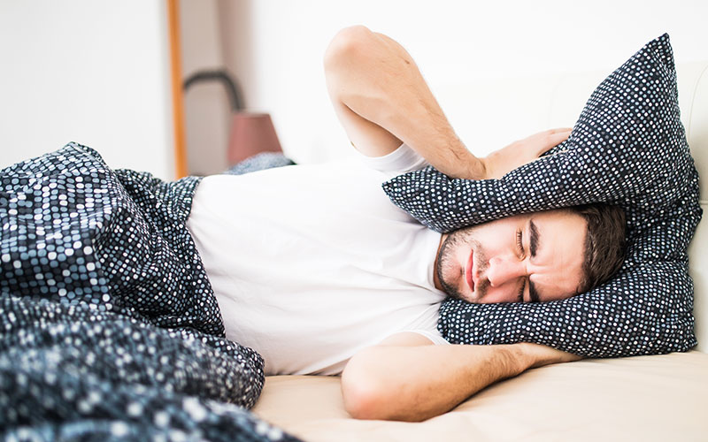 Handsome man covering his head and ears blocking out the sound with a pillow, suffering from loud alarm wake-up signal, young guy in bed awoken by a noise around him | Does Tinnitus Go Away | 9 Signs That Tinnitus is Going Away