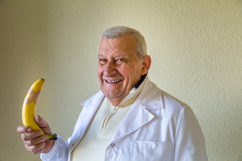 Old country doctor smiling while holding a large and erect banana with medical patch as concept of treating impotence | Erections