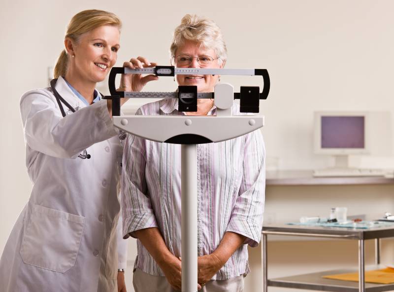 Doctor weighing senior woman in doctor office | Preventative Medicine