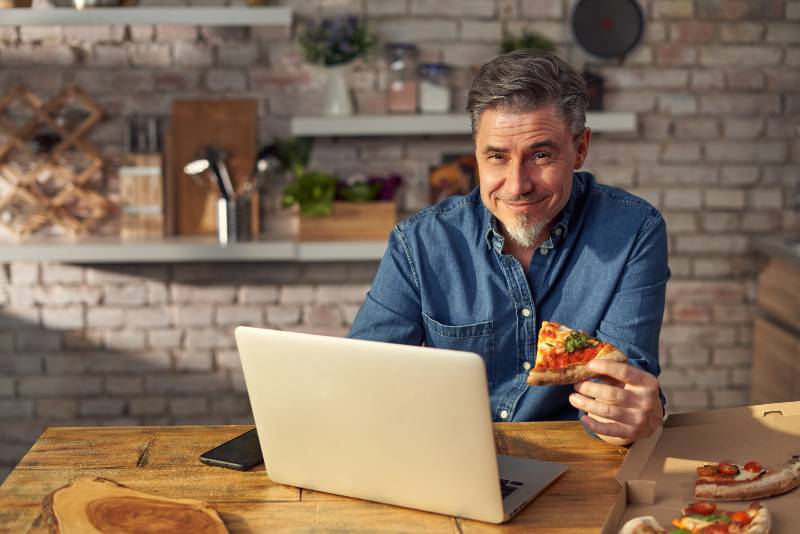 Man working from home on laptop computer, sitting at table in kitchen, eating online ordered pizza | Do Carbs Make You Fat