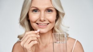 Smiling happy healthy middle aged 50s woman holding glass of water taking dietary supplement vitamin | Old women multivitamins antioxidants for anti age beauty | Calcium D Glucarate