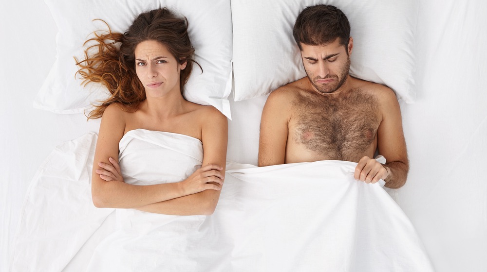 disappointed and depressed because of erectile dysfunction, looking at his penis under white cover, doesn't know what's the problem, angry wife lying next to him and waiting